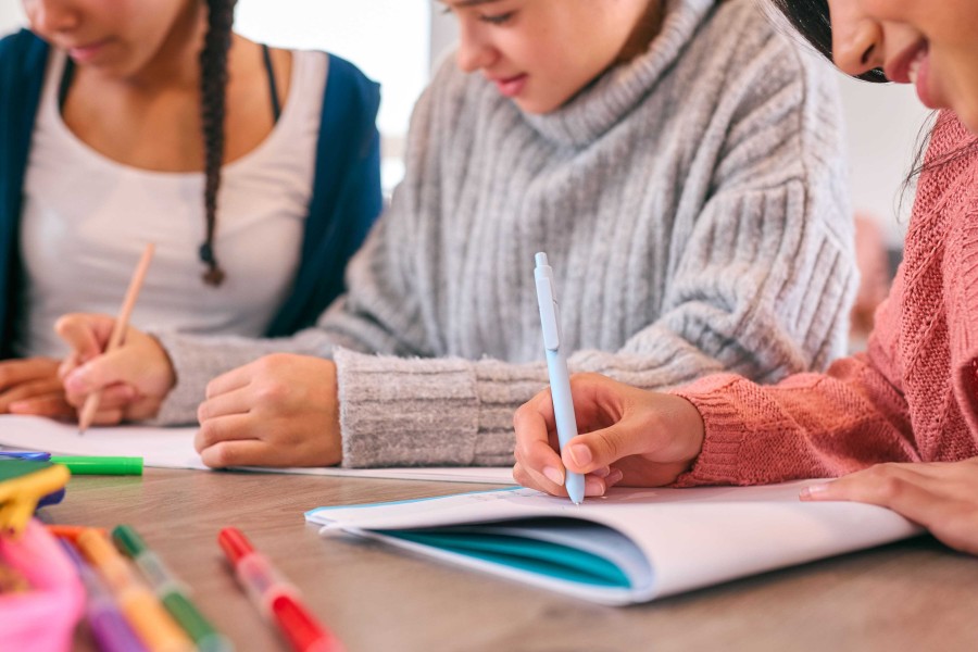 Three young women are seated at a wooden table, focused on their work, with notebooks, pens, and colored pencils scattered around.