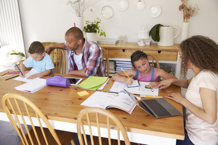 A family of four is gathered around a wooden dining table, engaged in homeschooling activities with books, laptops, and school supplies spread out.