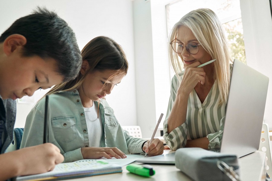 A teacher assisting two students with their work at a table with a laptop and school supplies.