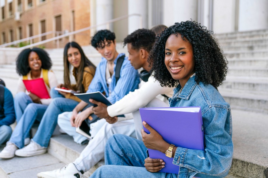 A diverse group of college students sitting on steps outside a university building, smiling and holding books and folders.