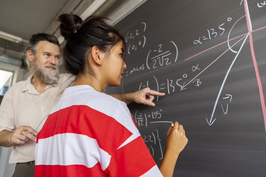A female high school student in a red and white striped shirt writing trigonometry equations on a chalkboard while her mature teacher observes.