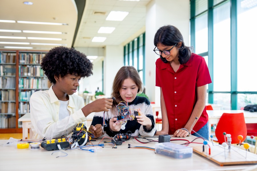 Three high school students assembling wiring on a small robot model at a desk in a spacious library setting.