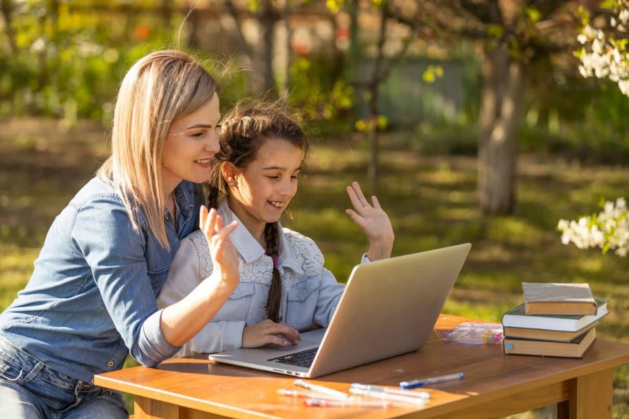 A mother and young daughter waving at a laptop screen while sitting together at a wooden table outdoors in a garden setting.