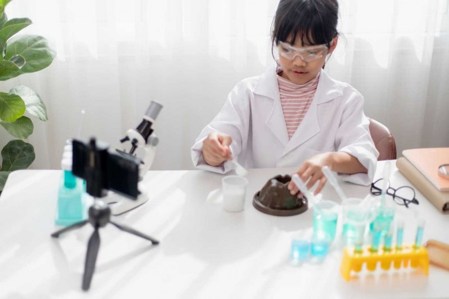 A young girl in a white lab coat and safety goggles recording a hands-on volcano science experiment using a smartphone mounted on a mini tripod.