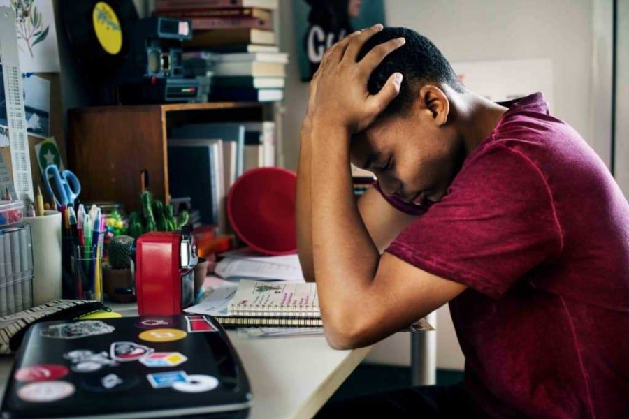 A teenage black male in a maroon t-shirt sitting at a cluttered desk with their head in their hands, representing student stress, burnout, and executive dysfunction.