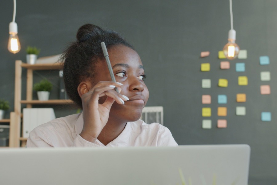 A young woman in a light-colored shirt holds a pen to her head while thinking at a desk in front of a laptop.