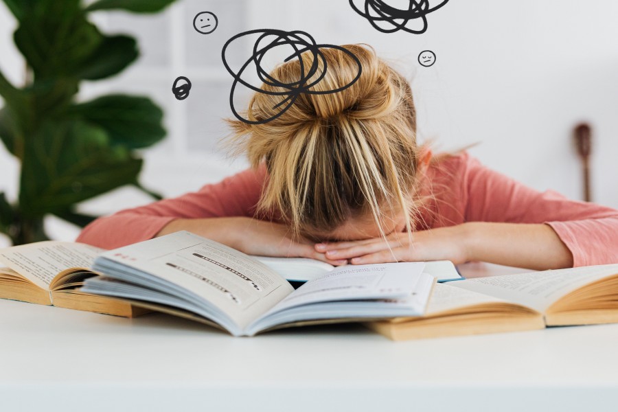 A girl in a pink shirt with blonde hair in a messy bun resting their face in their hands, slumped over open books at a table, surrounded by scribbled lines and stressed facial expression doodles.