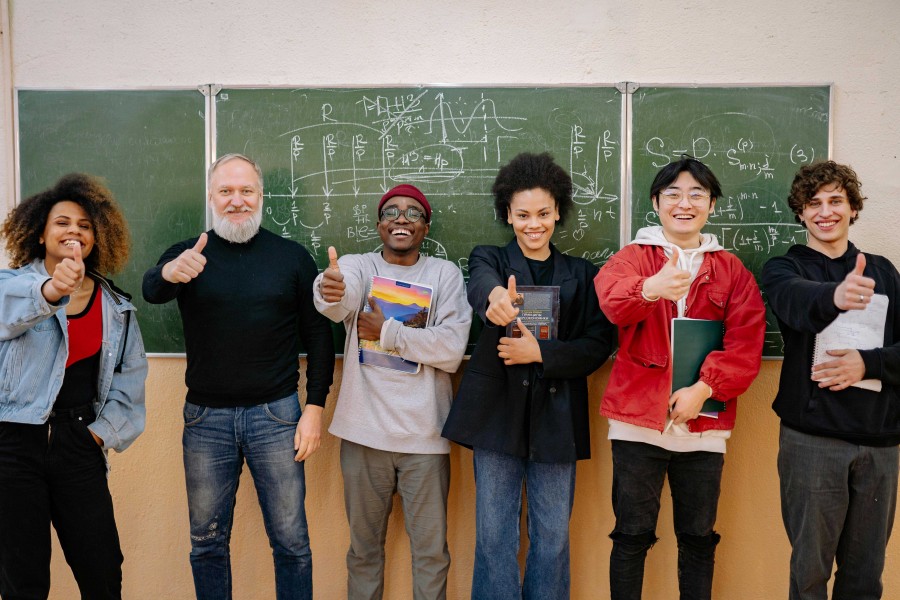 Diverse group of students and a teacher giving thumbs up in front of a chalkboard filled with math equations.