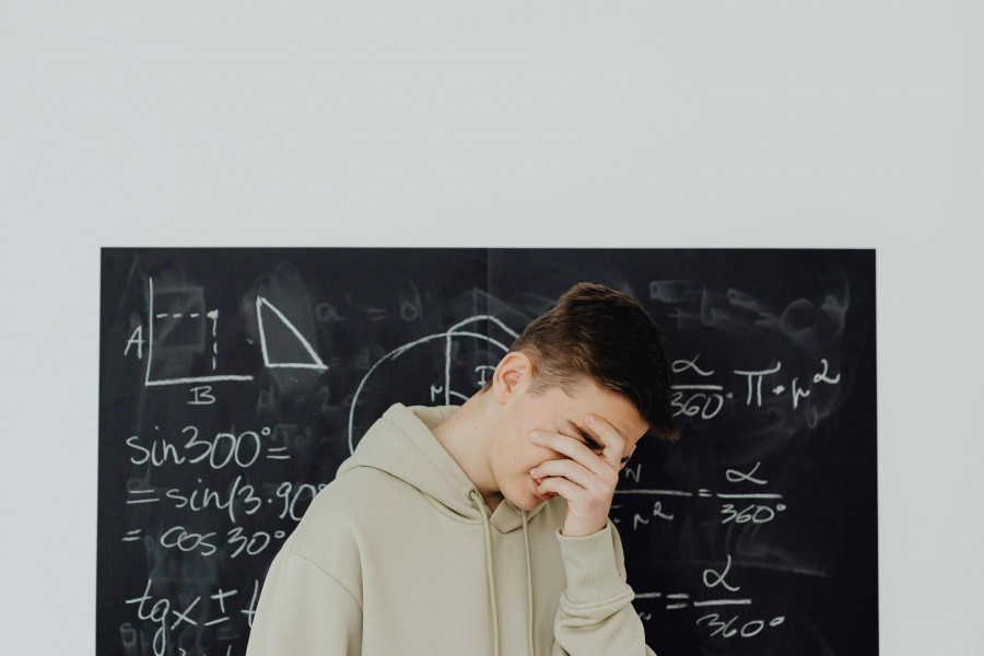 Young man in a beige hoodie standing in front of a blackboard covered in complex trigonometric and geometric math formulas.