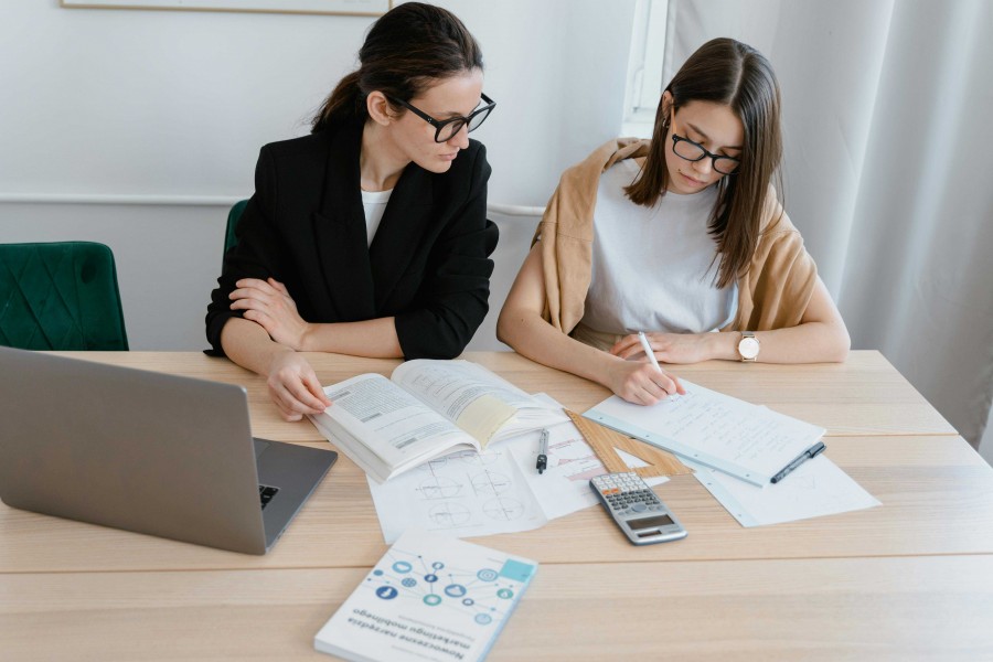 Two women at a wooden desk reviewing study materials. A tutor in a black blazer observes a student in a white shirt and tan