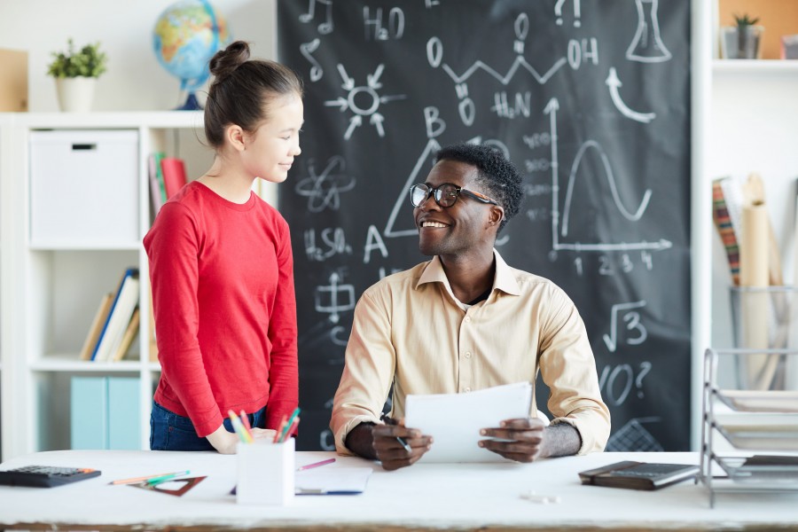 Young female student in red shirt smiles at a male teacher wearing glasses, seated at a desk covered in papers and a calculator.