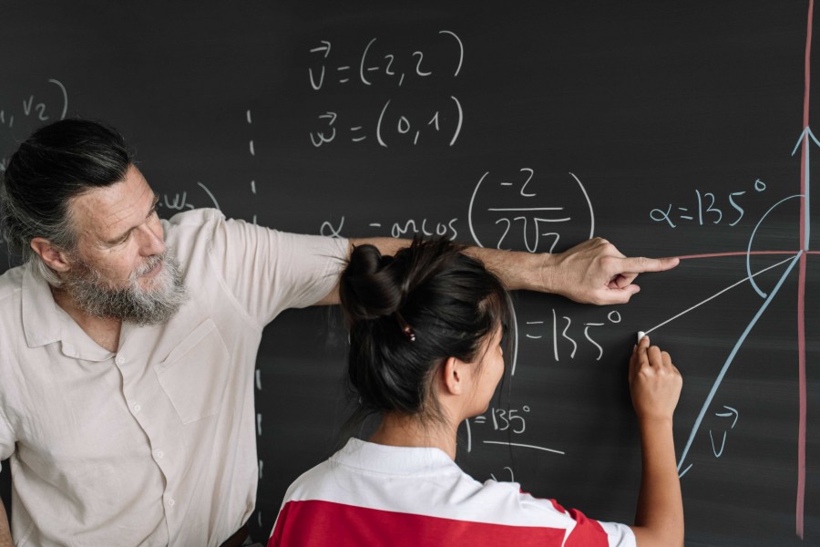 A middle-aged male teacher with a gray beard and light shirt points at a blackboard covered with mathematical equations and vector notations, while an Asian female student, viewed from behind, writes a solution with chalk.